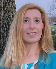 A woman with long blonde hair, blue eyes, and a blue and white striped shirt standing outdoors near a tree, with houses and a cloudy sky in the background.