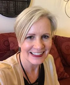 A smiling woman with short blonde hair taking a selfie indoors, sitting on a red-patterned sofa.