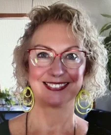 A woman with curly blonde hair wearing glasses and large yellow hoop earrings, smiling in an indoor setting.