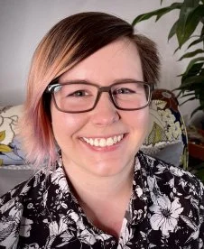 A woman with short hair wearing glasses and a floral shirt, smiling indoors with a plant in the background.