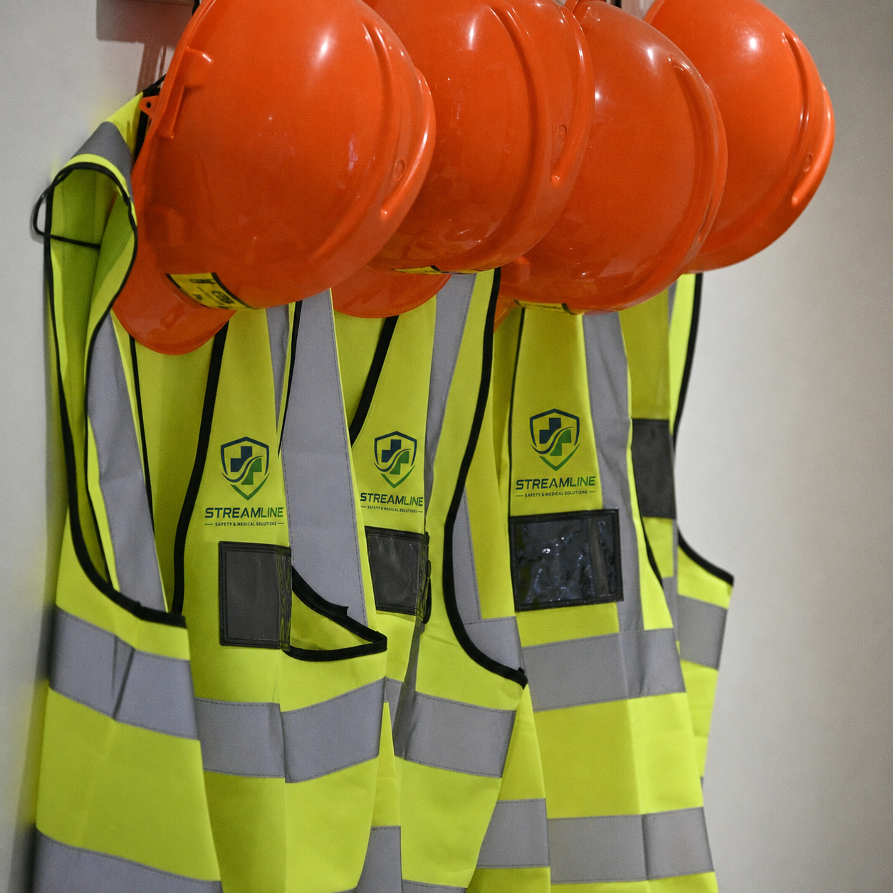Several high-visibility safety vests with reflective stripes hanging on a wall, each with an orange safety helmet resting on top, marked with the logo and name 'Streamline'.