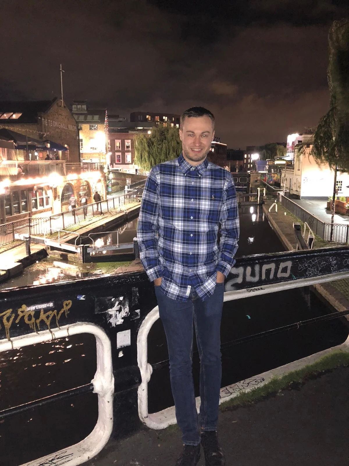 A man in a blue plaid shirt and jeans standing outdoors at night near a canal with city buildings and lights in the background.