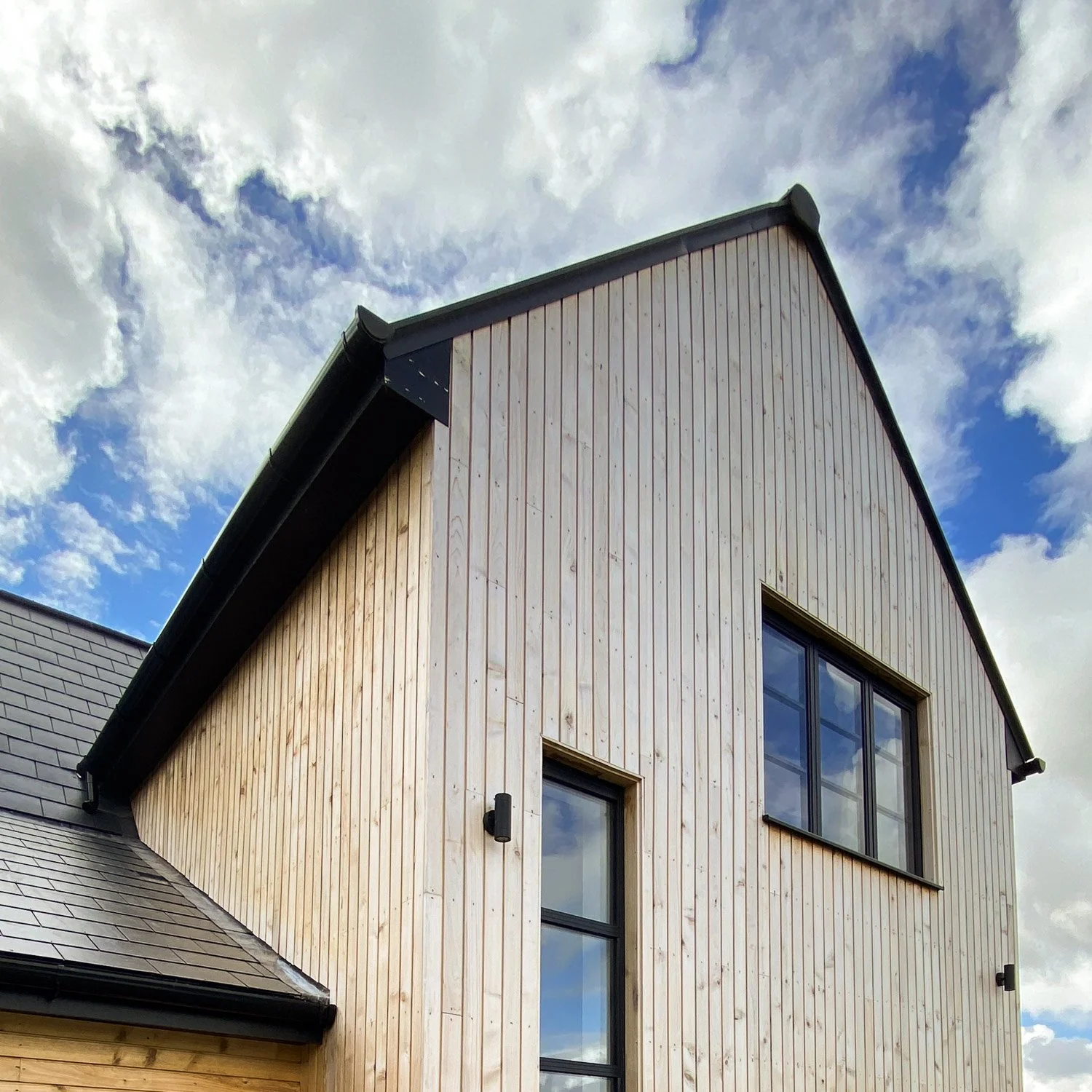 Close-up of a house exterior with vertical wood siding and large window, under a cloudy sky.