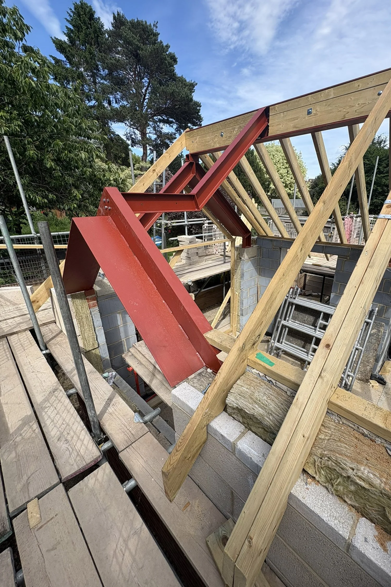 Construction site showing wooden and metal framework for a building, with a partially built roof structure and scaffolding, surrounded by trees and blue sky.