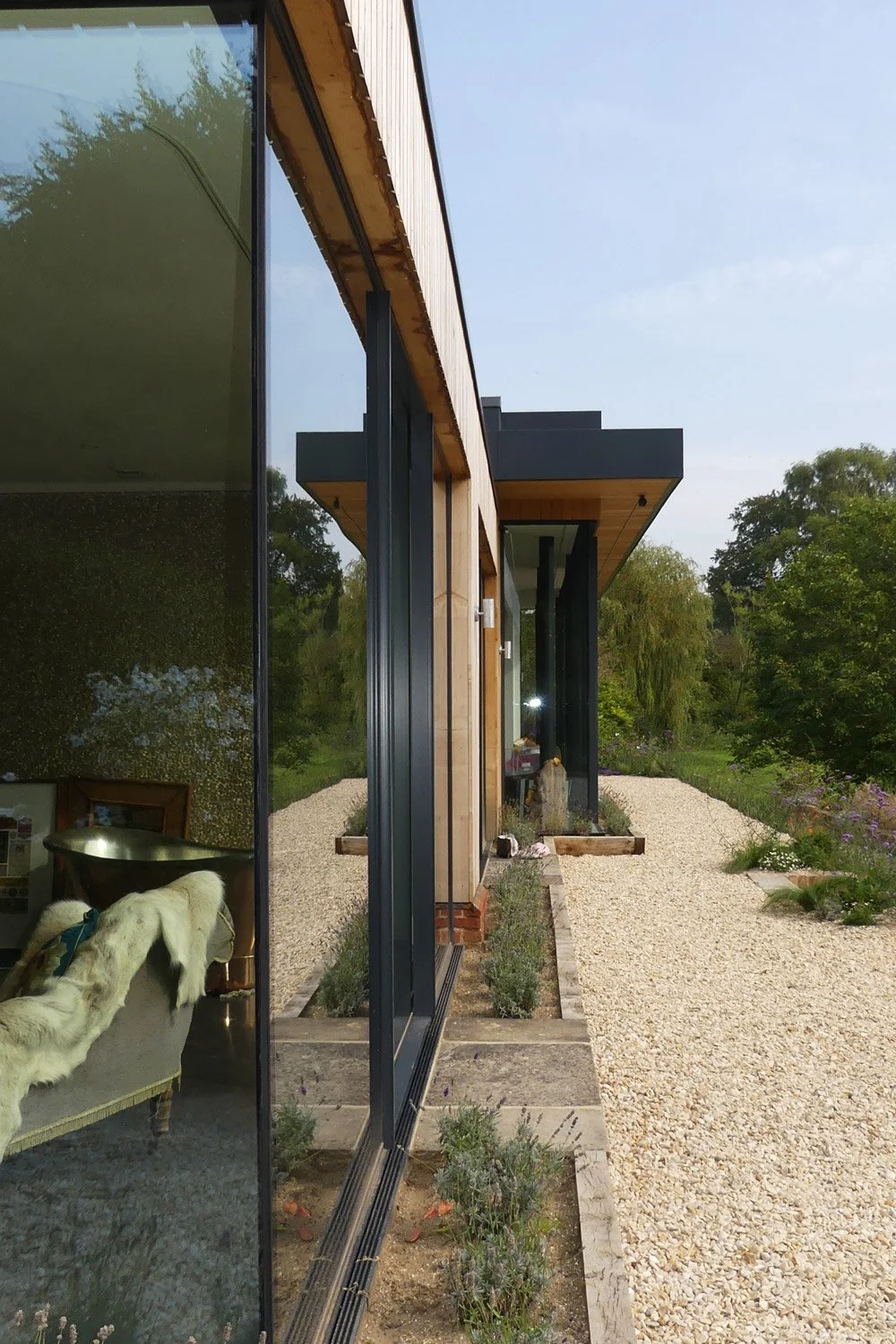 Side view of a modern house with large glass windows, wooden exterior, and landscaped gravel pathway surrounded by greenery.