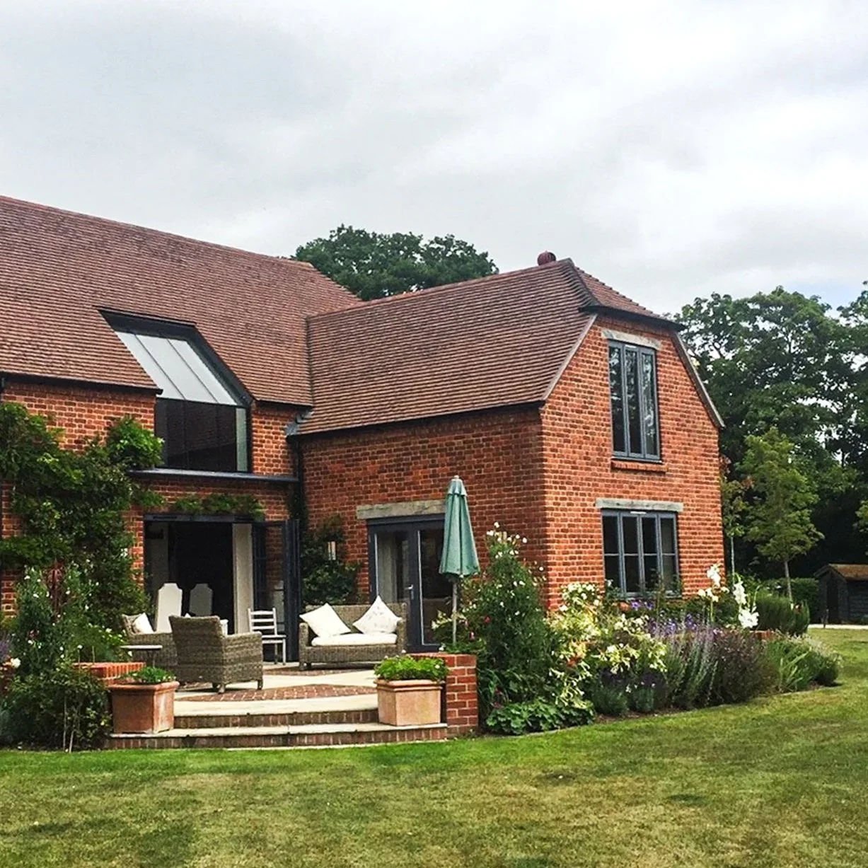 Red brick house with a patio, outdoor furniture, potted plants, and a garden with flowers, under an overcast sky.