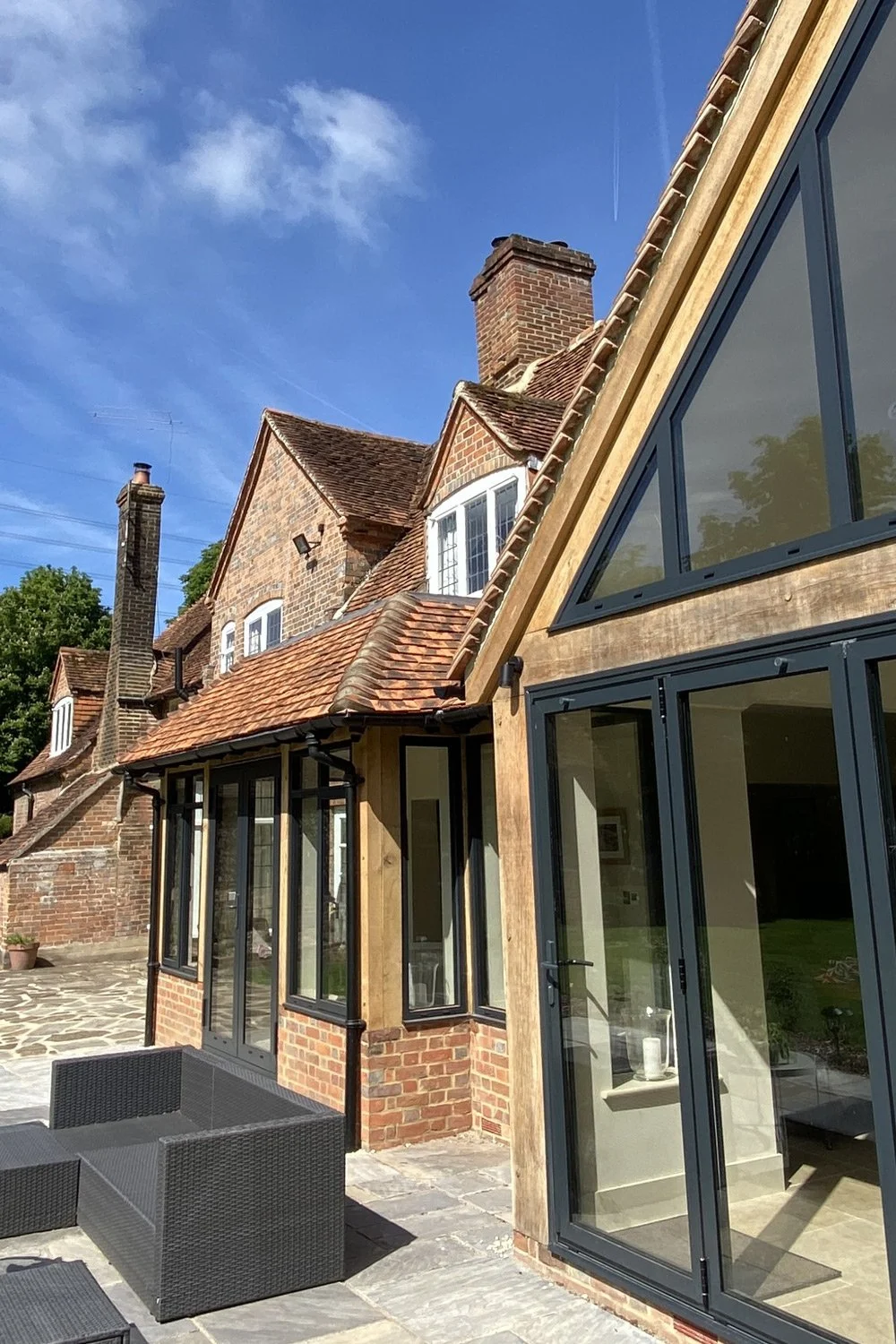 Old brick house with traditional roof and chimneys, adjacent modern extension with large glass windows and wooden accents, outdoor patio with black wicker furniture under a blue sky.