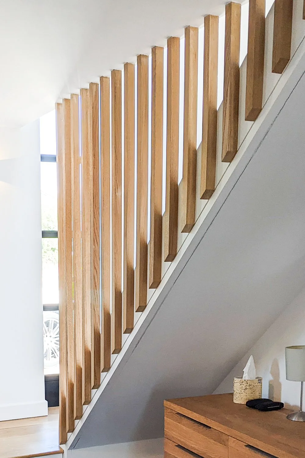 Interior view of a staircase with vertical wooden railings and a wooden dresser below, next to a wall with a lamp and tissue box.