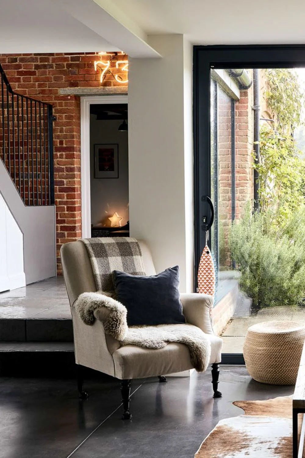Living room with a beige armchair, dark cushion, and cozy throw blanket, near a glass door leading outside, exposed brick wall, modern decor, sunlight streaming in, and a round woven ottoman.