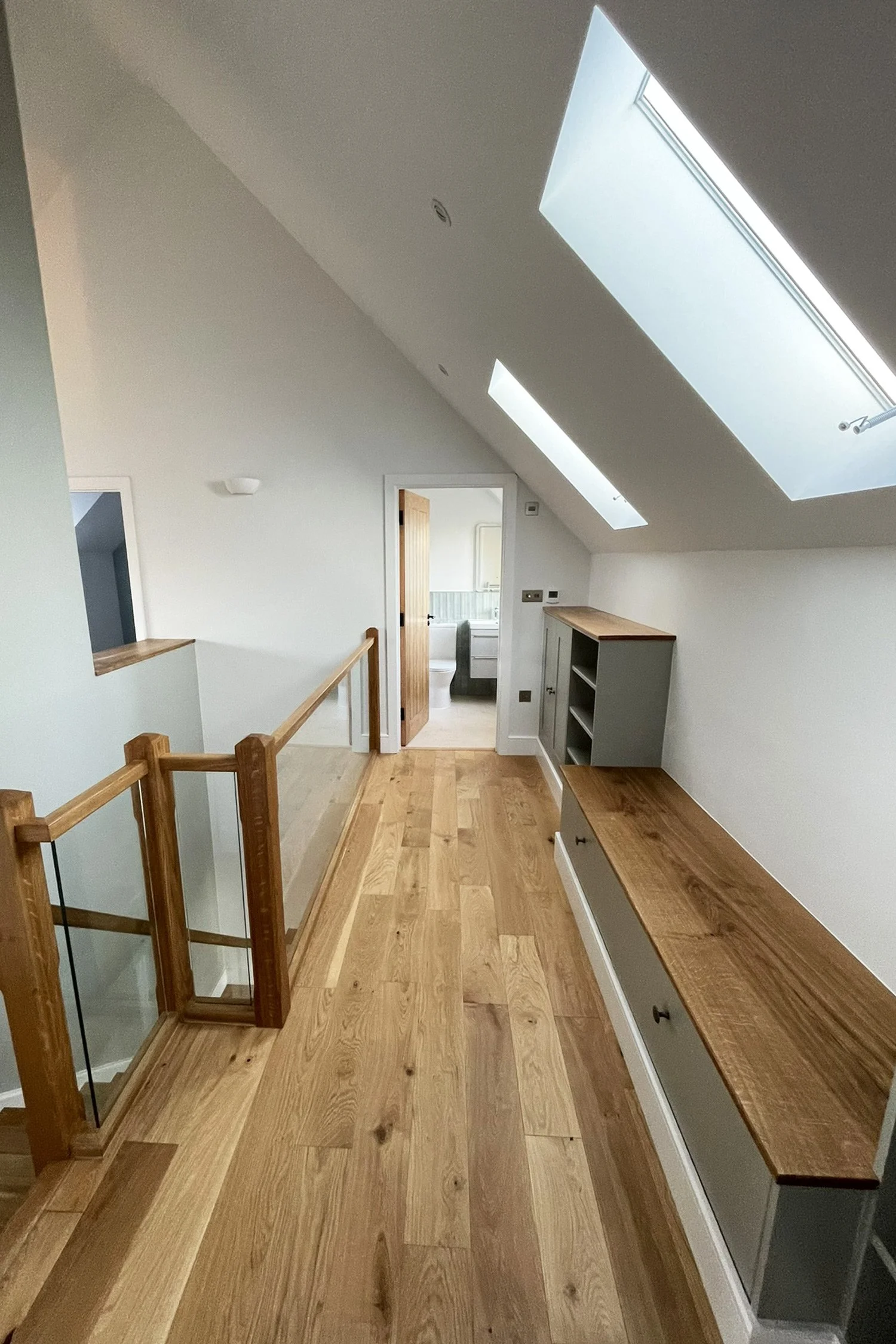 Interior view of an attic space with sloped ceiling, skylights, wooden flooring, a small gray shelving unit, and a bathroom visible through an open door.