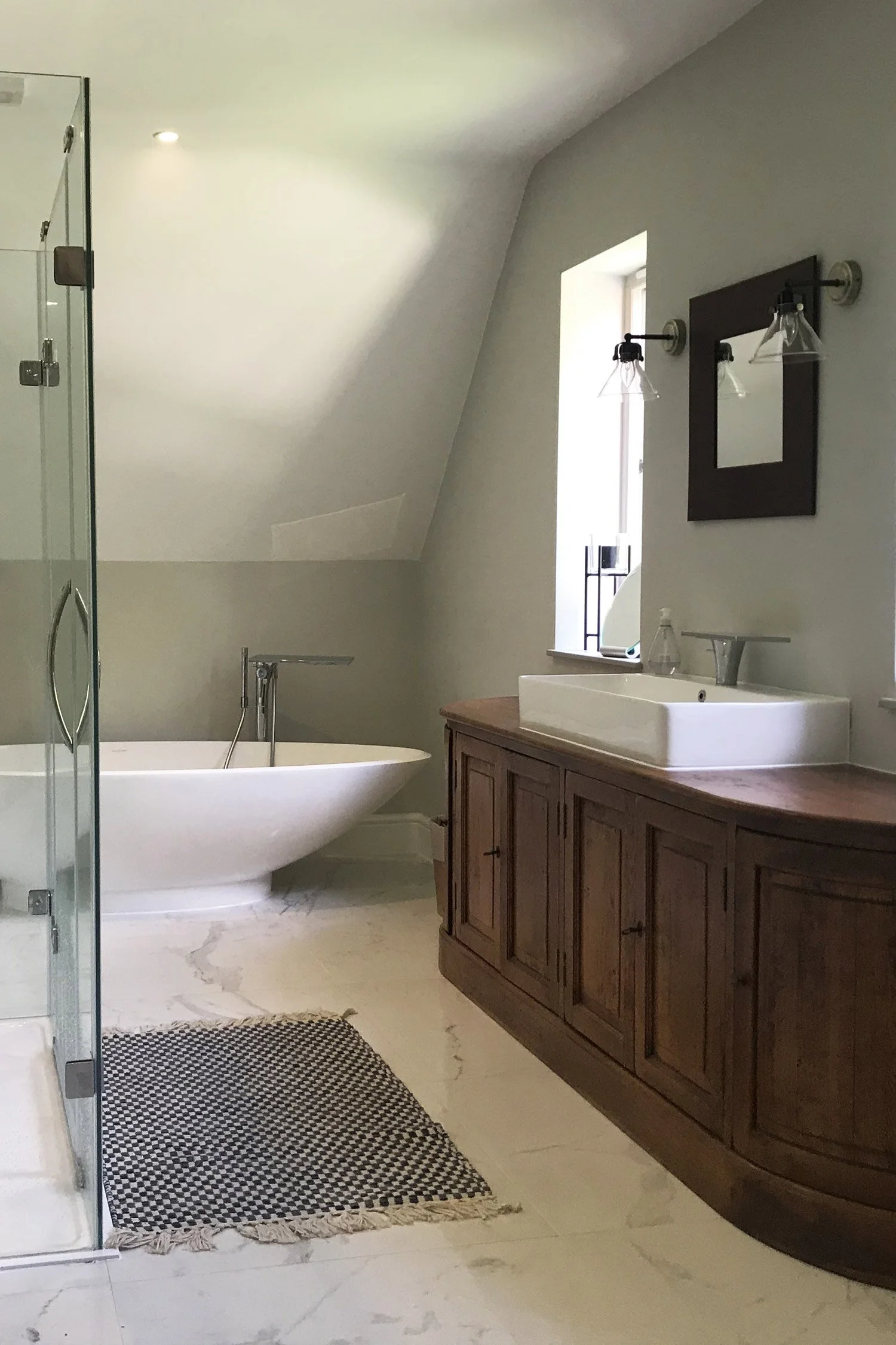 Bathroom with a bathtub, wooden vanity with a sink, mirror, wall-mounted lights, window, and a shower enclosure with glass door. There is a black and white patterned rug on the marble floor.