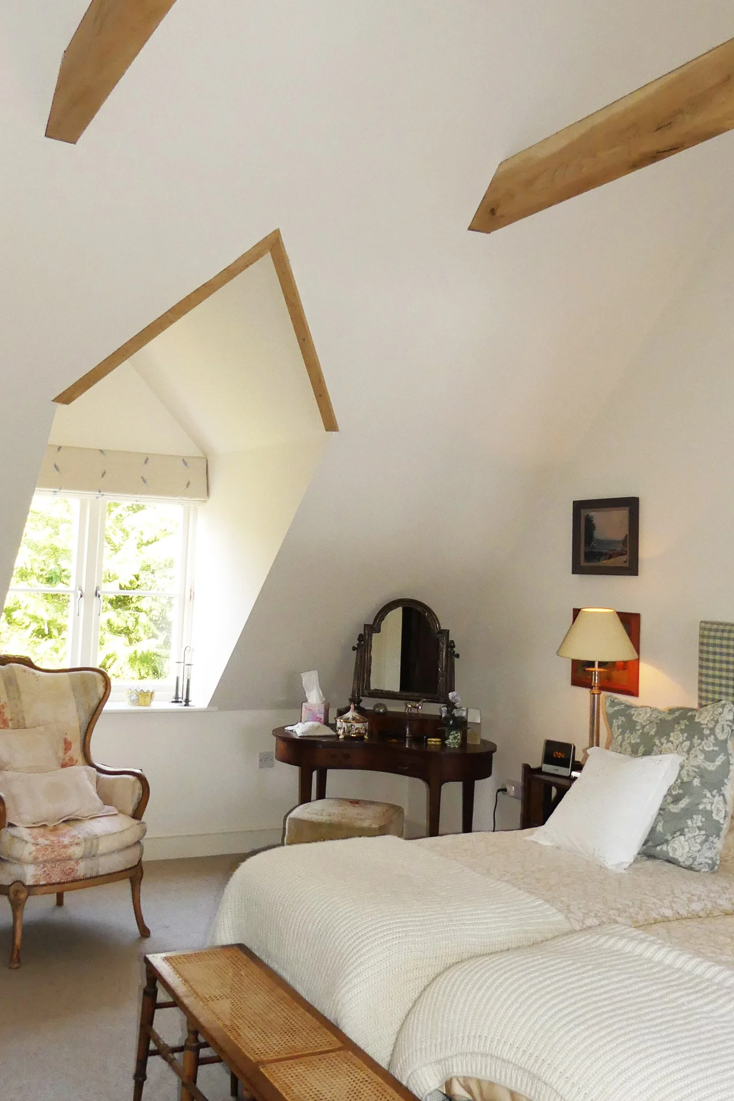 A cozy bedroom with a sloped ceiling and wooden beams. There is a large window letting in natural light with a white curtain. The room has vintage-style furniture including an armchair, a dresser with a mirror, and a bed with floral pillow and white 