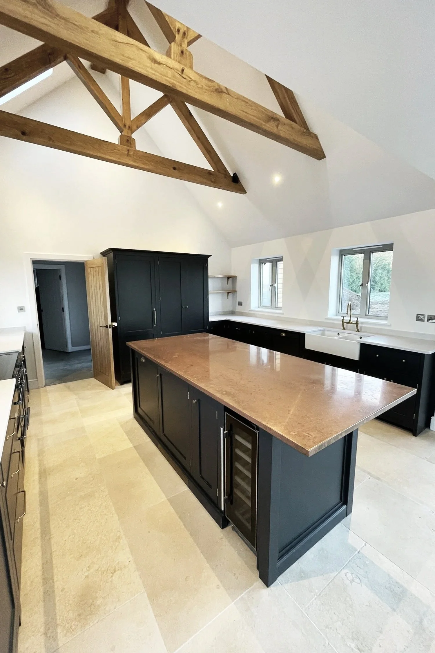 Modern kitchen with black cabinetry, a large central island with a brown countertop, white walls, and three windows above the sink. Exposed wooden beams on the ceiling and light-colored tiled flooring.