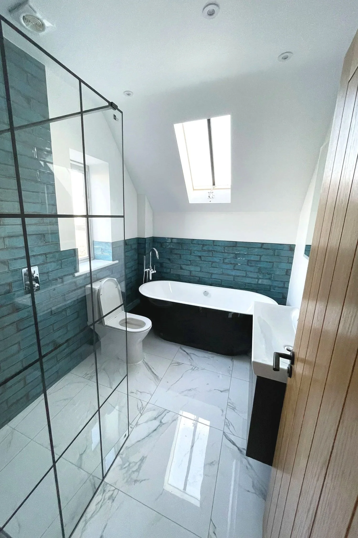 Modern bathroom with white marble floor, a freestanding bathtub, a small wall-mounted sink, a toilet, and a window. The room has a skylight, wooden door, and a partial glass-enclosed shower area with black metal framing and greenish-blue brick wall a