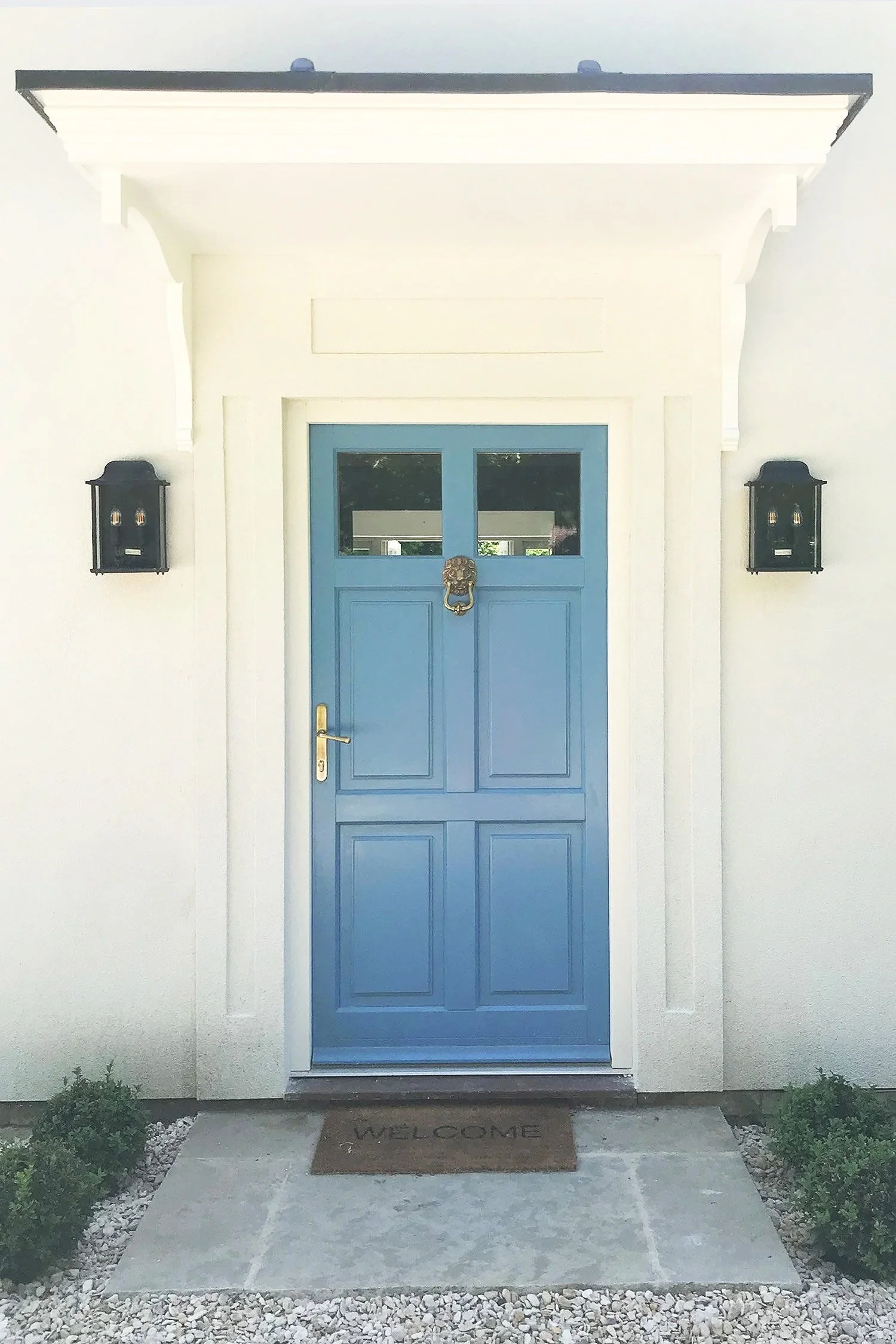 Blue front door with two glass panels, a brass handle, and a lion head door knocker, flanked by black wall-mounted lanterns, with a concrete walkway, white pebbled borders, and a welcome mat.