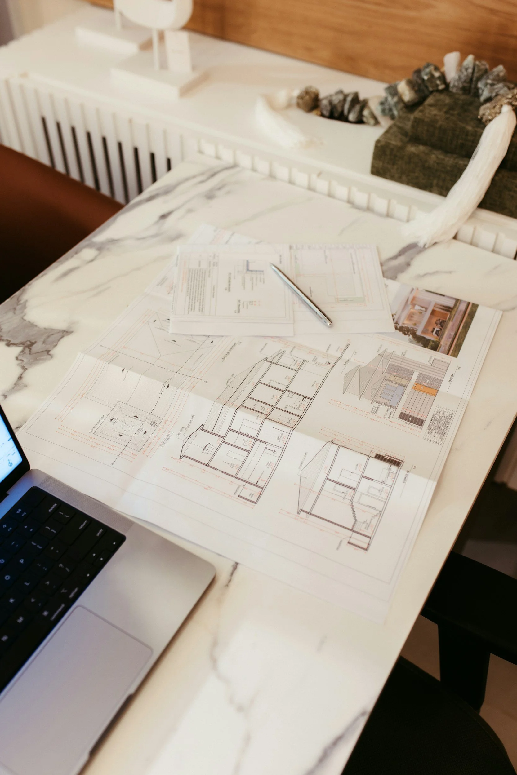 Architectural blueprints and drawings spread on a marble table with a laptop and pen, with a headboard and rocks in the background.