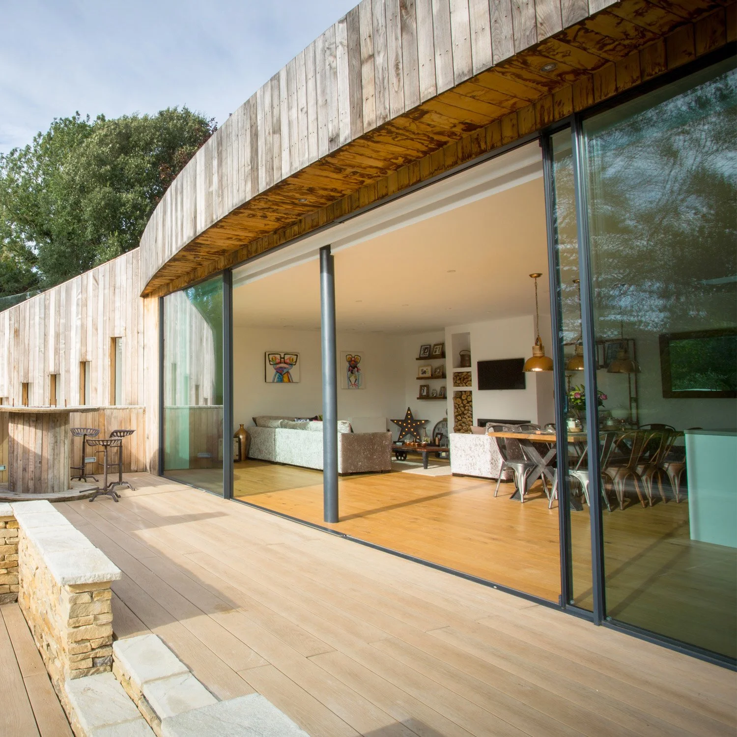 Modern house with large glass sliding doors revealing a living room and dining area inside. Exterior features wooden and stone elements, with a small balcony and outdoor seating. Trees are visible behind the house.