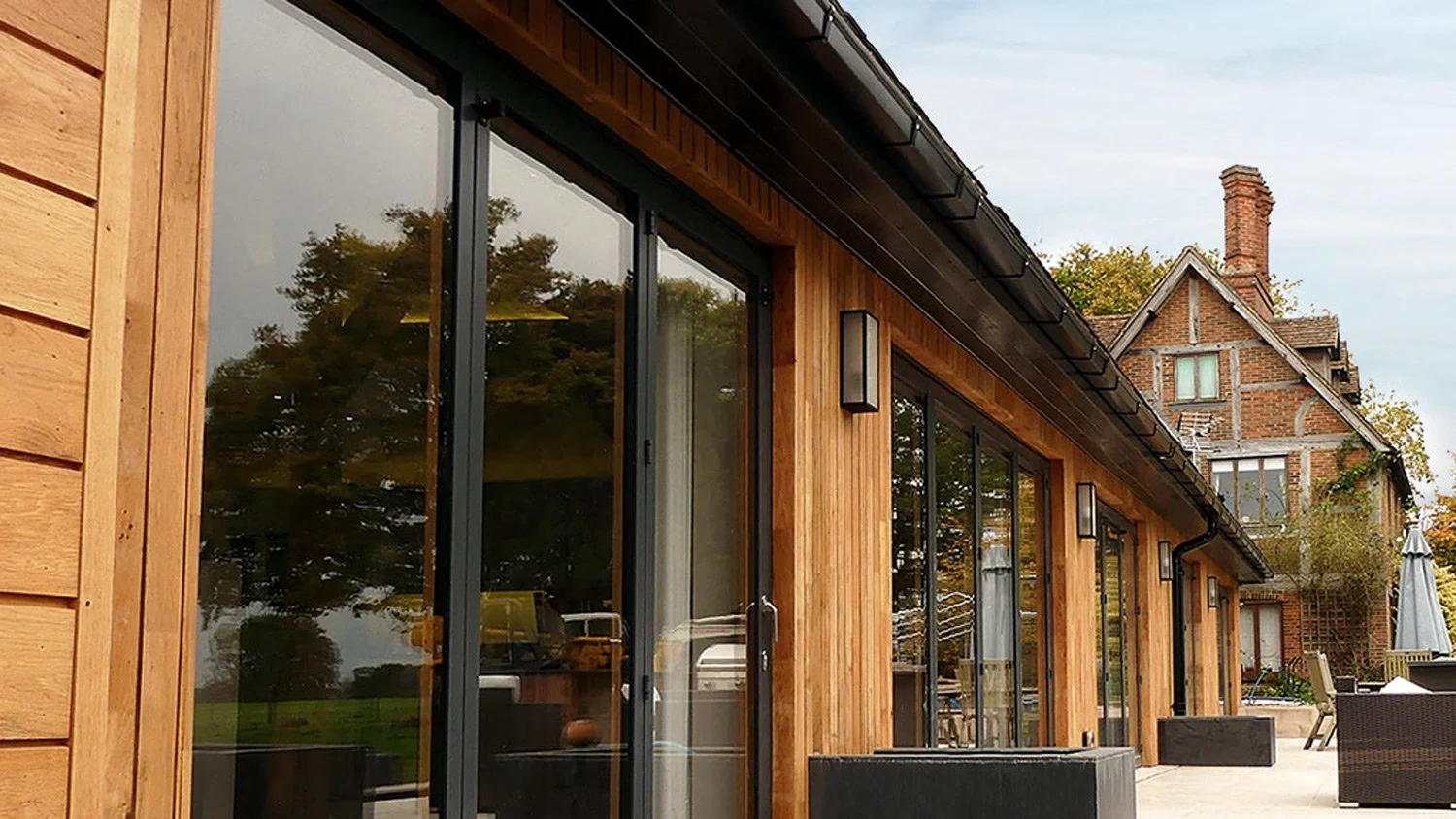 Modern house exterior with large glass sliding doors, wooden siding, outdoor furniture, and older brick house in background.