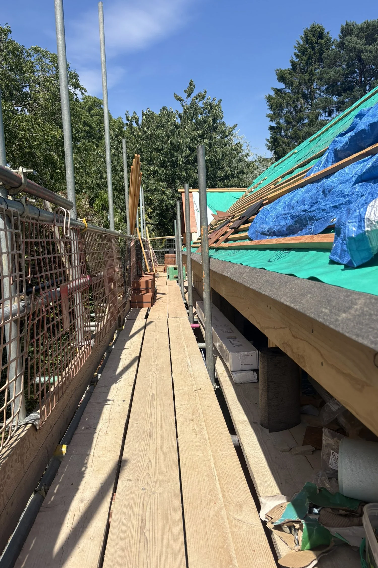 Construction site with wooden planks laid over scaffolding, building materials, and a partially completed roof with green and blue tarps, surrounded by trees and a clear blue sky.