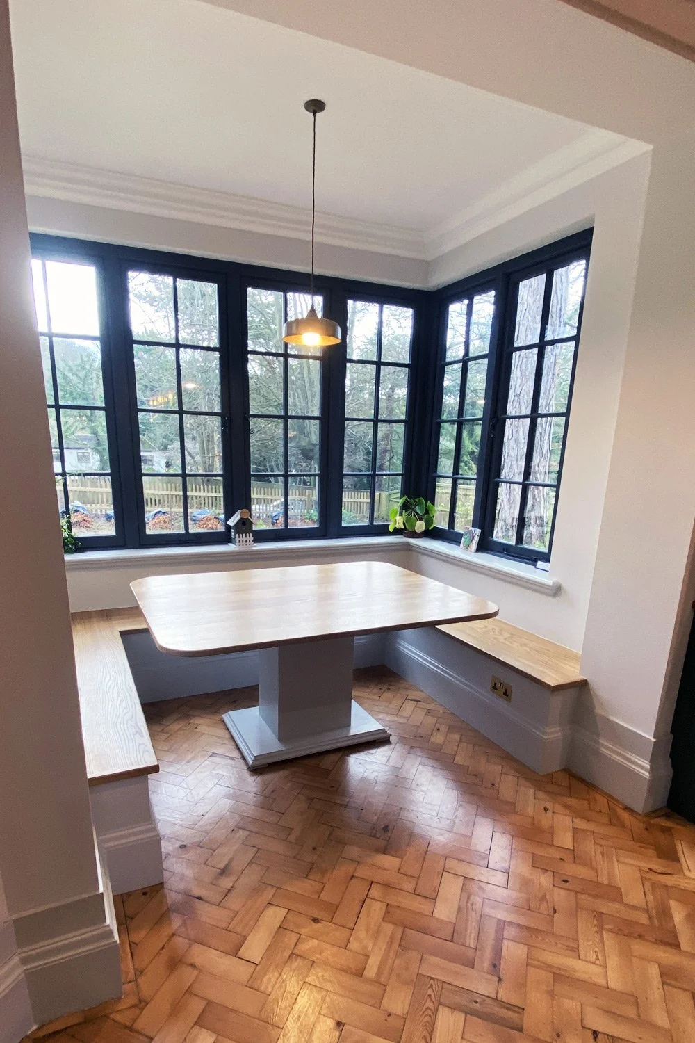 A cozy corner with a wooden table, built-in bench seating, large black-framed windows, hardwood herringbone flooring, a hanging light fixture, and decorative plants.