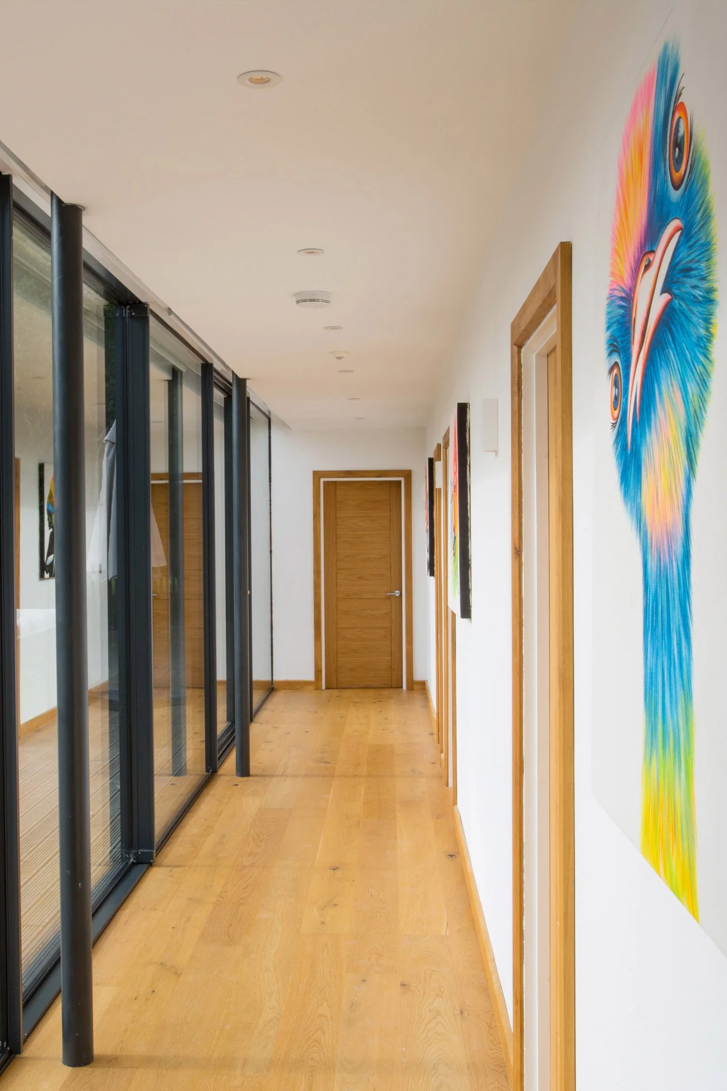 A hallway with wooden flooring, white walls decorated with colorful art, and large glass sliding doors with black frames on the left side.