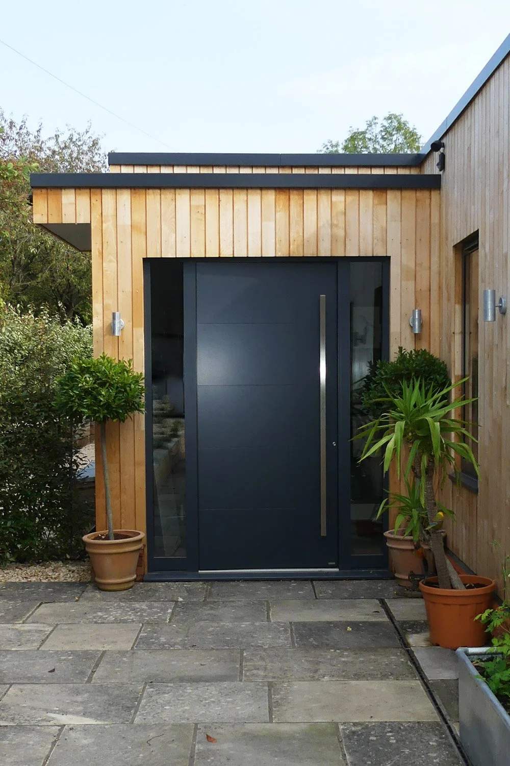 Modern house entrance with black door, wooden panel walls, potted plants, and stone pavement.