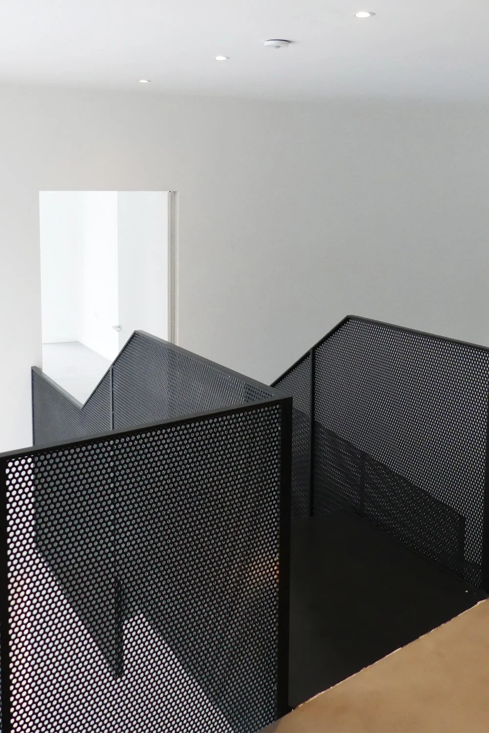 Modern black metal mesh stair railings in a minimalist interior with white walls and a doorway leading to bright room.