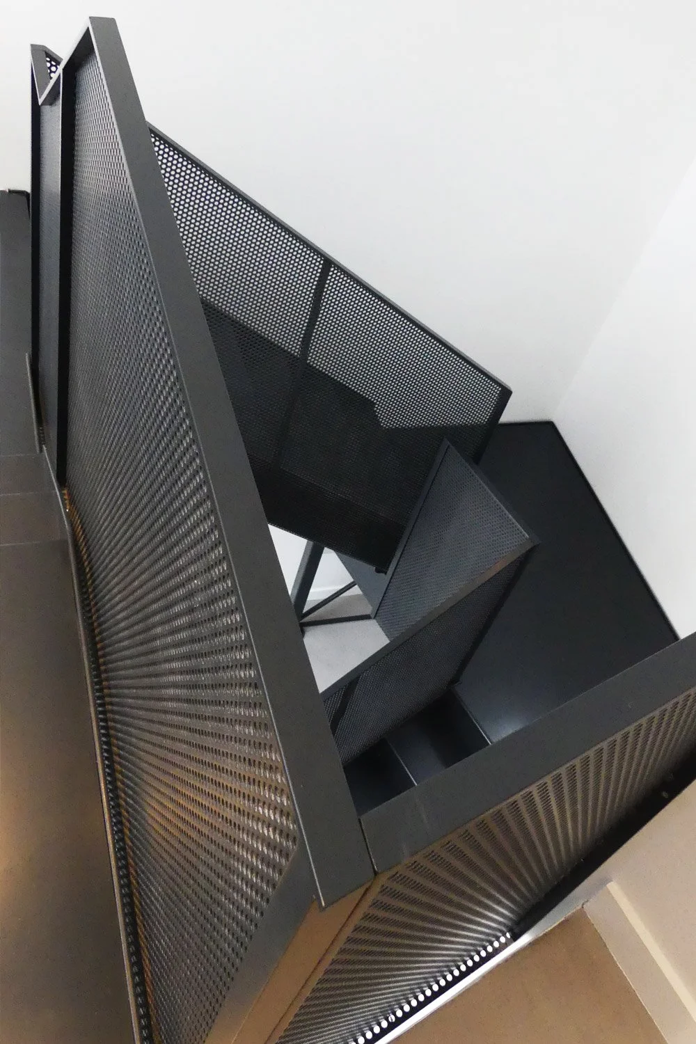 Top-down view of black perforated metal furniture or equipment with angular surfaces against a white wall.