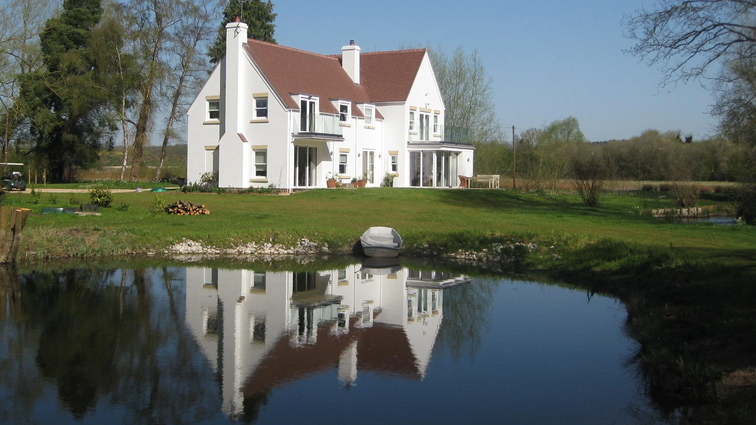 A white house with a red roof by a pond, with trees and grass surrounding it.
