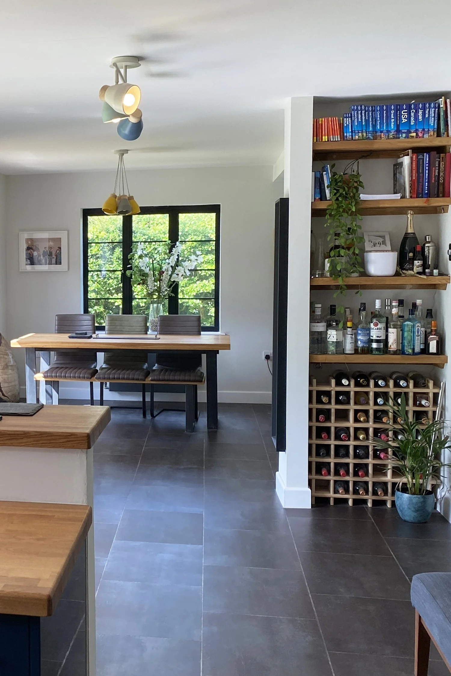 A dining area with a wooden table and gray chairs positioned in front of a window with green foliage outside. There's a multicolored ceiling light fixture and a built-in shelf filled with books, a potted plant, framed pictures, and liquor bottles, wi