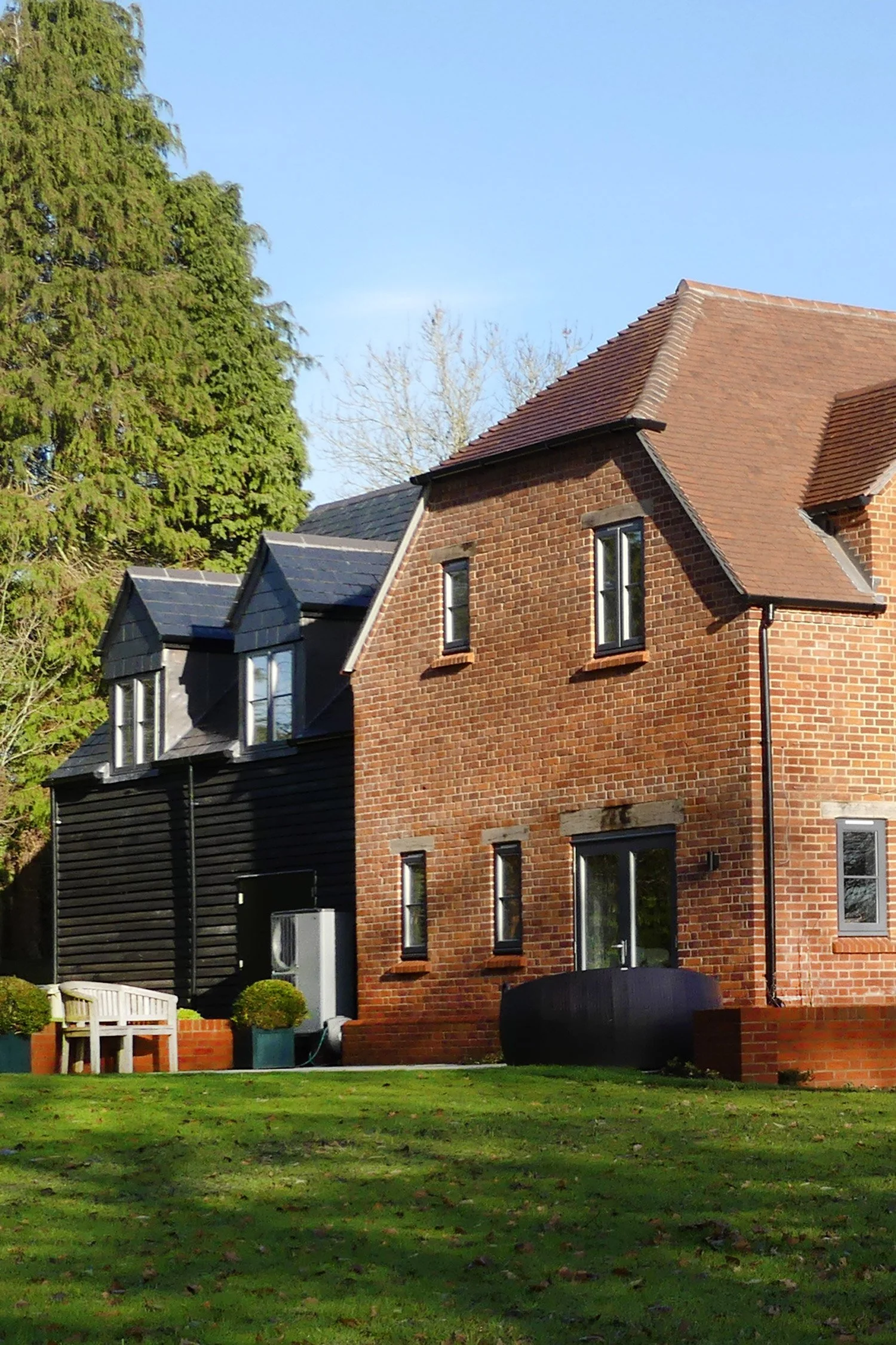 A brick house with a tiled roof and black window frames, surrounded by greenery, including a large tree and a grassy yard with a white bench and potted plants.