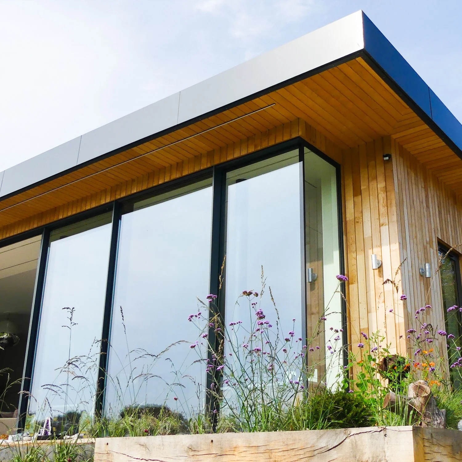 Modern house with large glass windows, wooden accents, and a flat roof, surrounded by wildflowers and greenery.