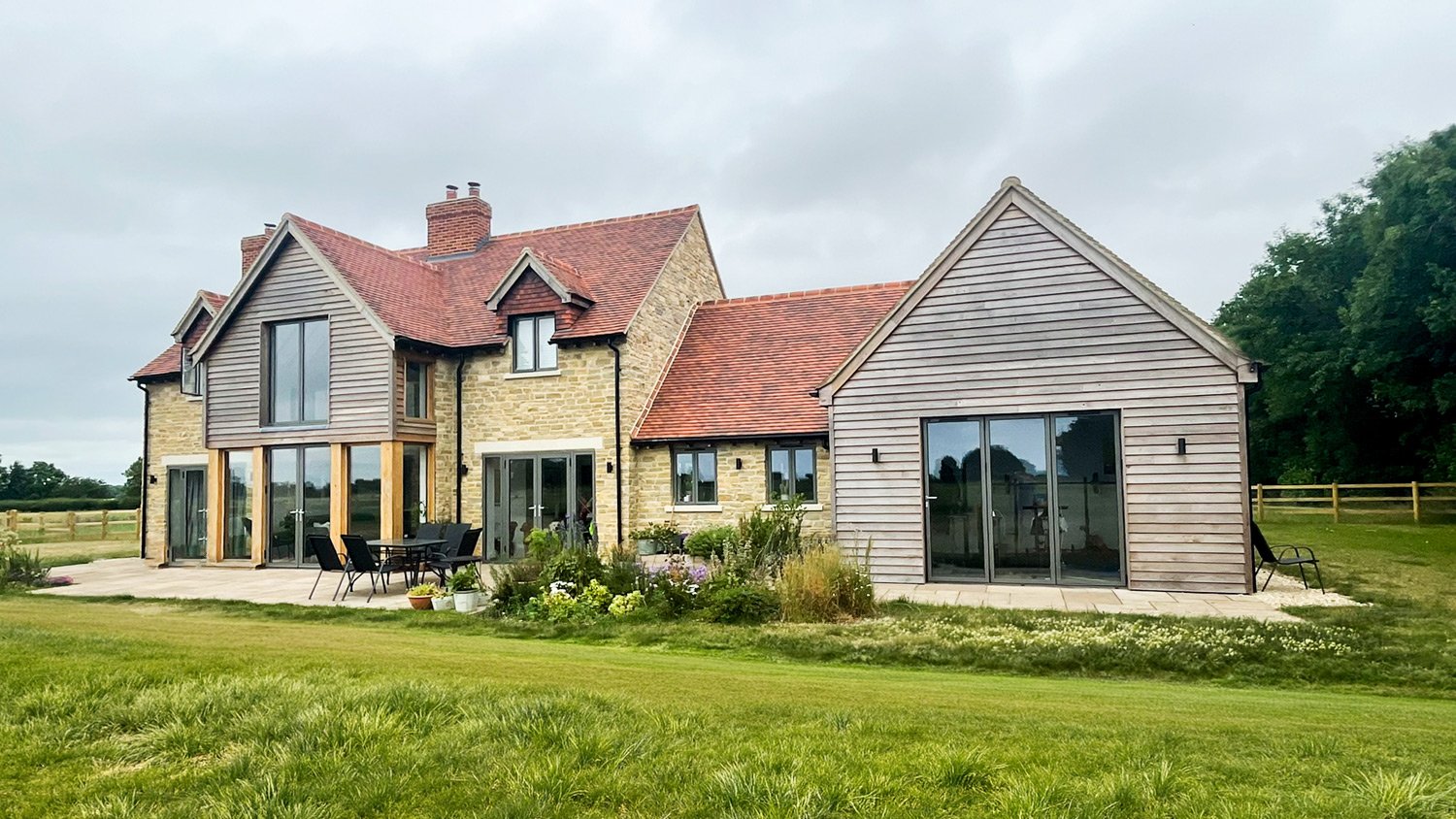A large house with brick and wooden siding, multiple gabled roofs with red tiles, and large glass sliding doors, surrounded by a green lawn and a patio with outdoor furniture.