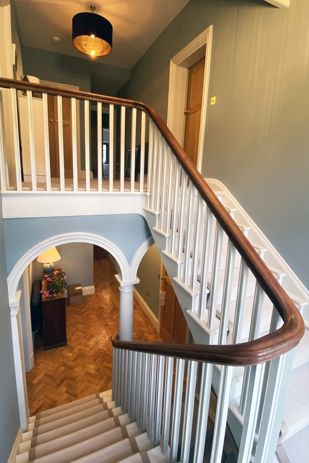 Interior view of a staircase inside a house with wooden and white railing, leading to a lower floor with hardwood flooring, a small table with flowers, and an arched doorway.