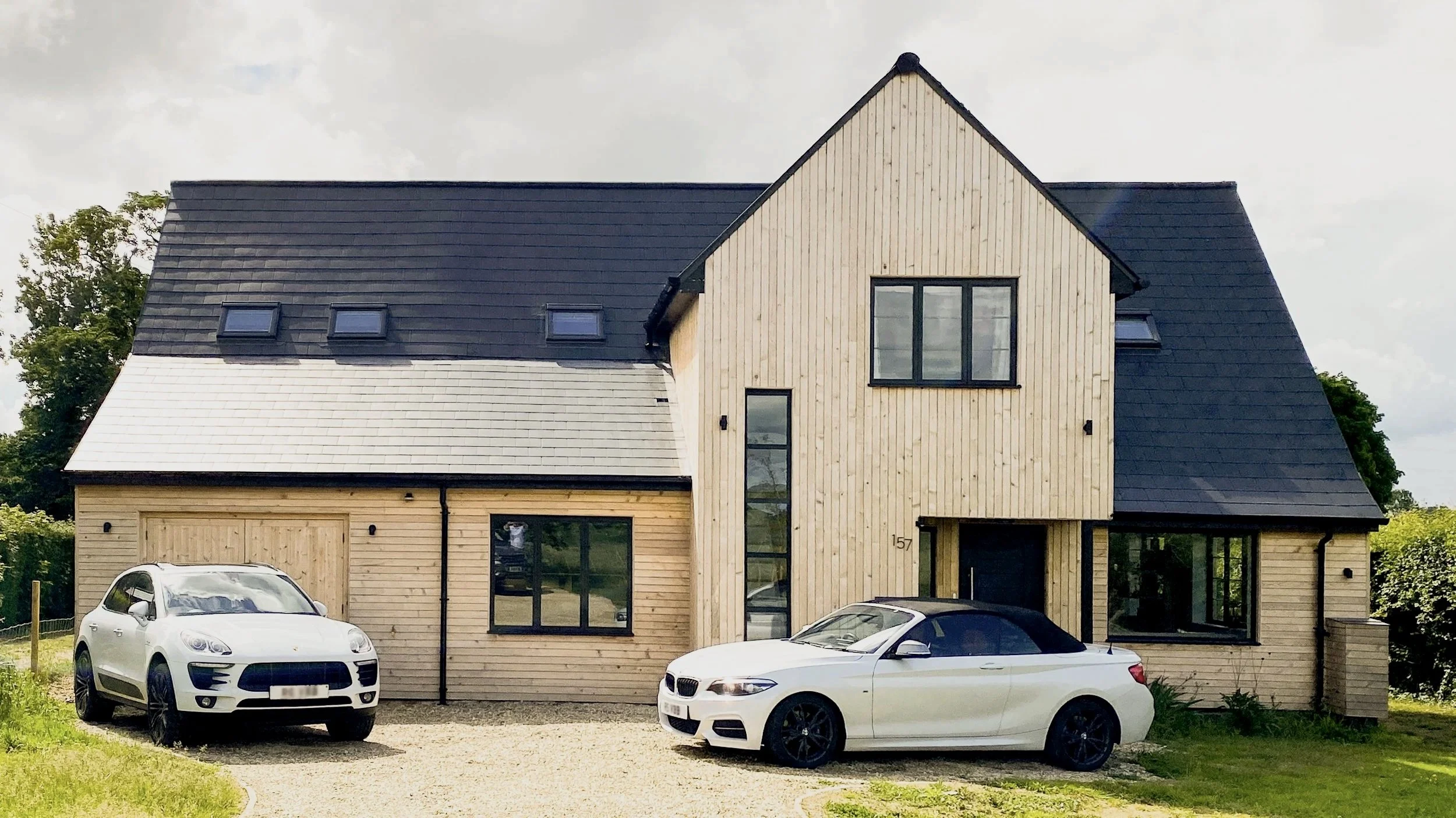 Modern house with a wooden exterior, black roof, and large windows, with three cars parked in front and greenery around.