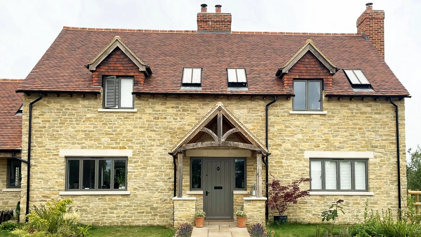 Front view of a two-story house with a brick and stone exterior, red tiled roof, dormer windows, and a small porch with a wooden gabled roof over the front door, surrounded by greenery and plants.