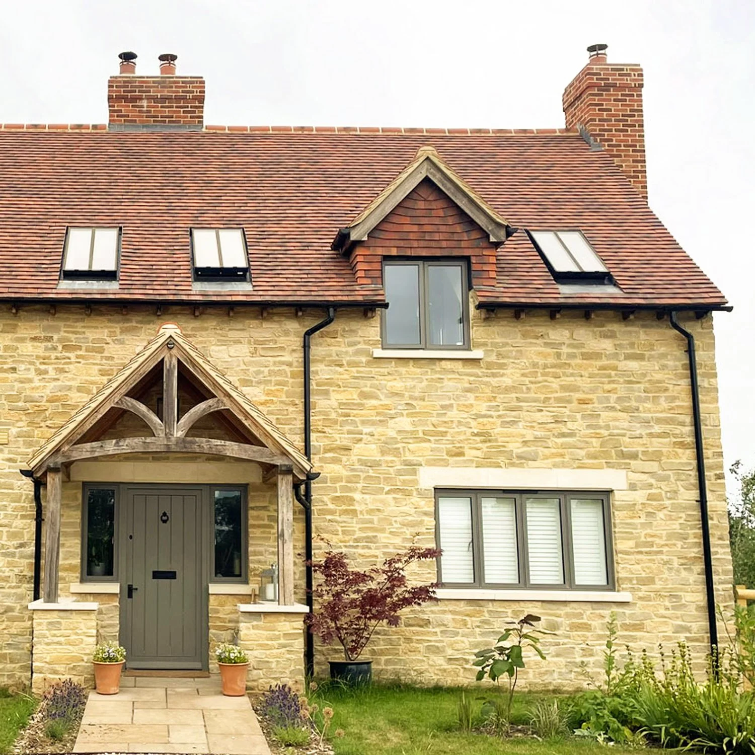 Front view of a stone house with a gabled roof, skylights, and two chimneys. The entrance has a small porch with wooden beams and a gray door, surrounded by flower pots and garden plants.