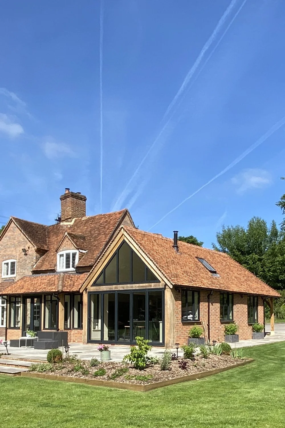 A brick house with a red tiled roof and large glass windows, surrounded by a well-maintained lawn and garden, under a blue sky with visible contrail streaks.
