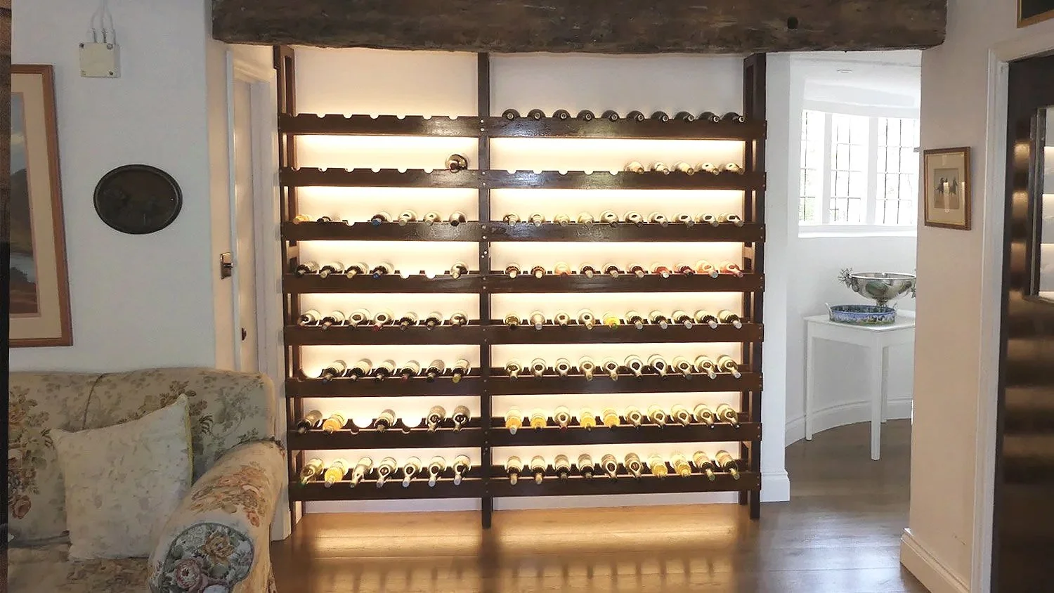 A wooden wine rack with multiple shelves holding numerous bottles of wine, placed against a white wall in a cozy, well-lit room.