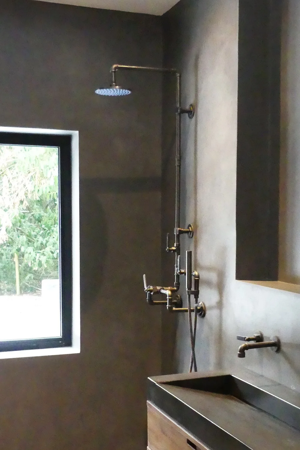 Indoor shower with multiple showerheads, a dark wall, a small window showing greenery outside, and a black sink with a wall-mounted faucet.