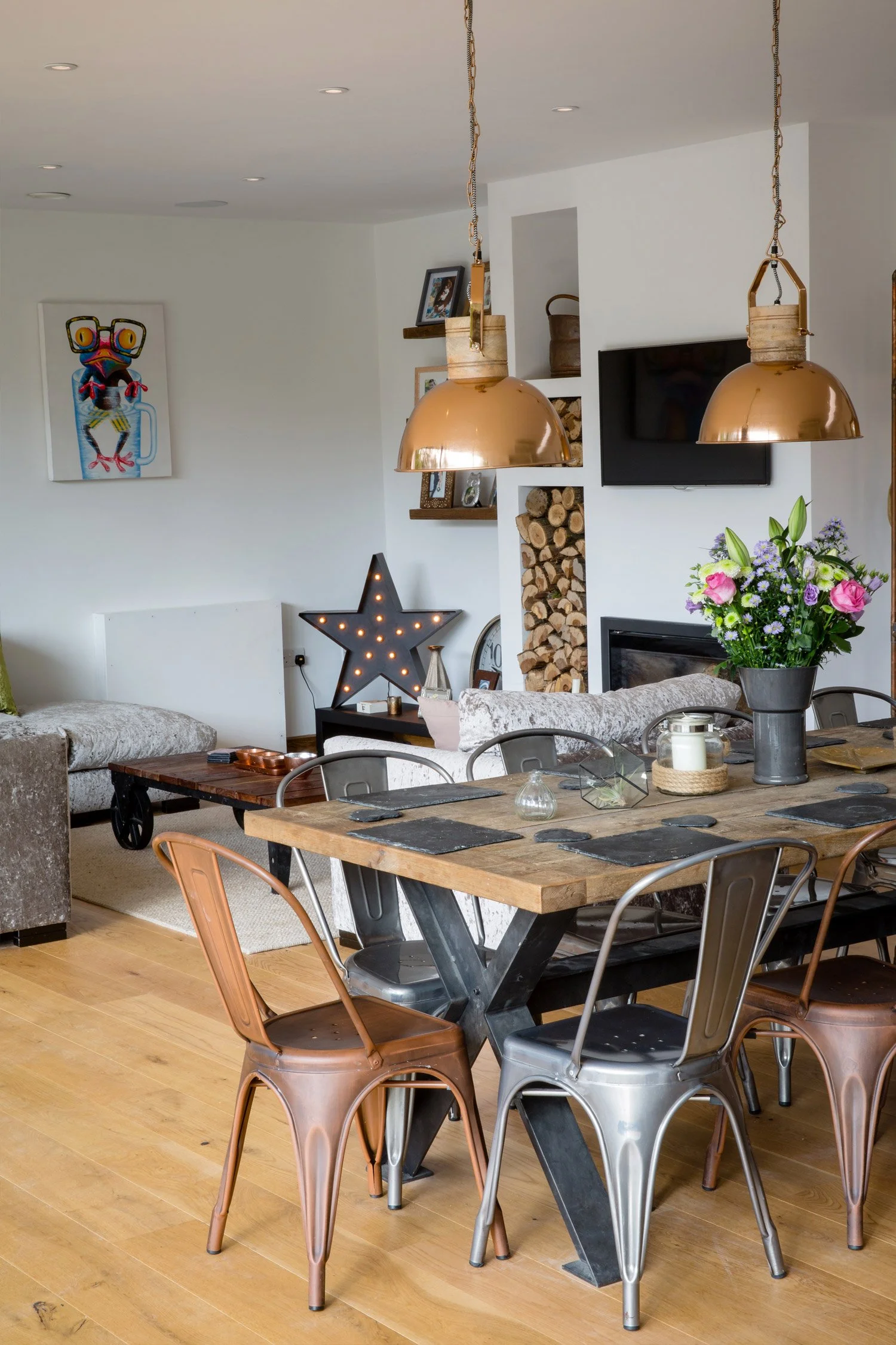 A living room with a wooden dining table, metal chairs, a gray sofa, decorative items like a star-shaped light, flowers in a vase, wall art and shelving, and a TV on the wall.