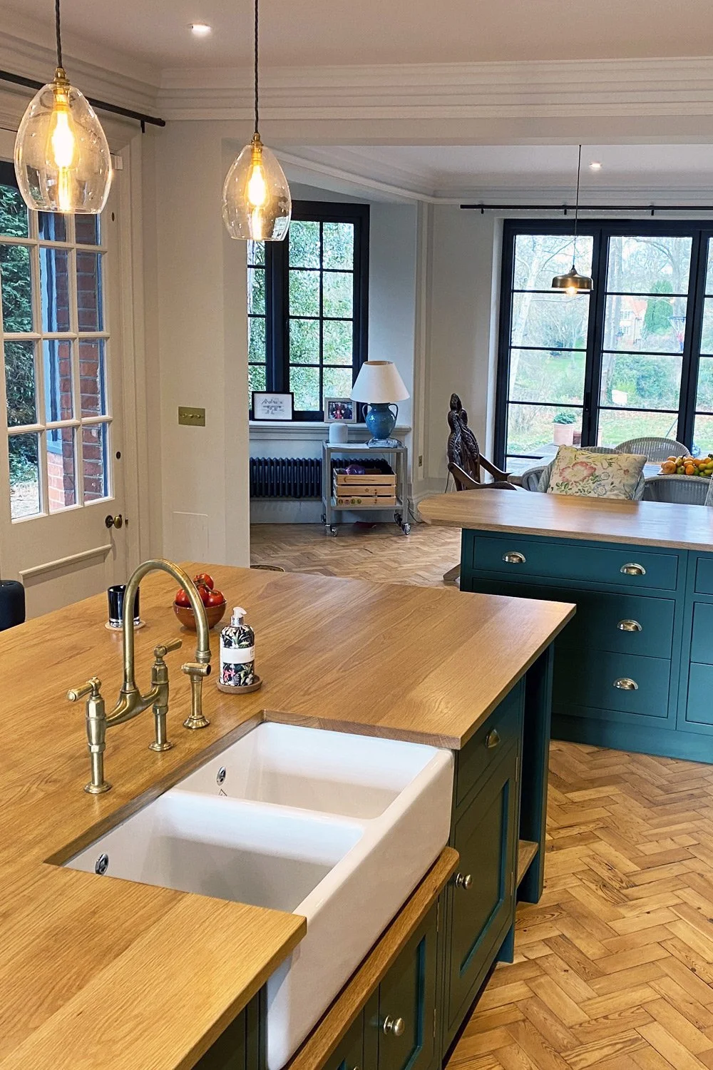 A modern kitchen with wooden countertops, white farmhouse sink, brass faucet, green cabinets, pendant lights, and large windows with a view of the backyard.