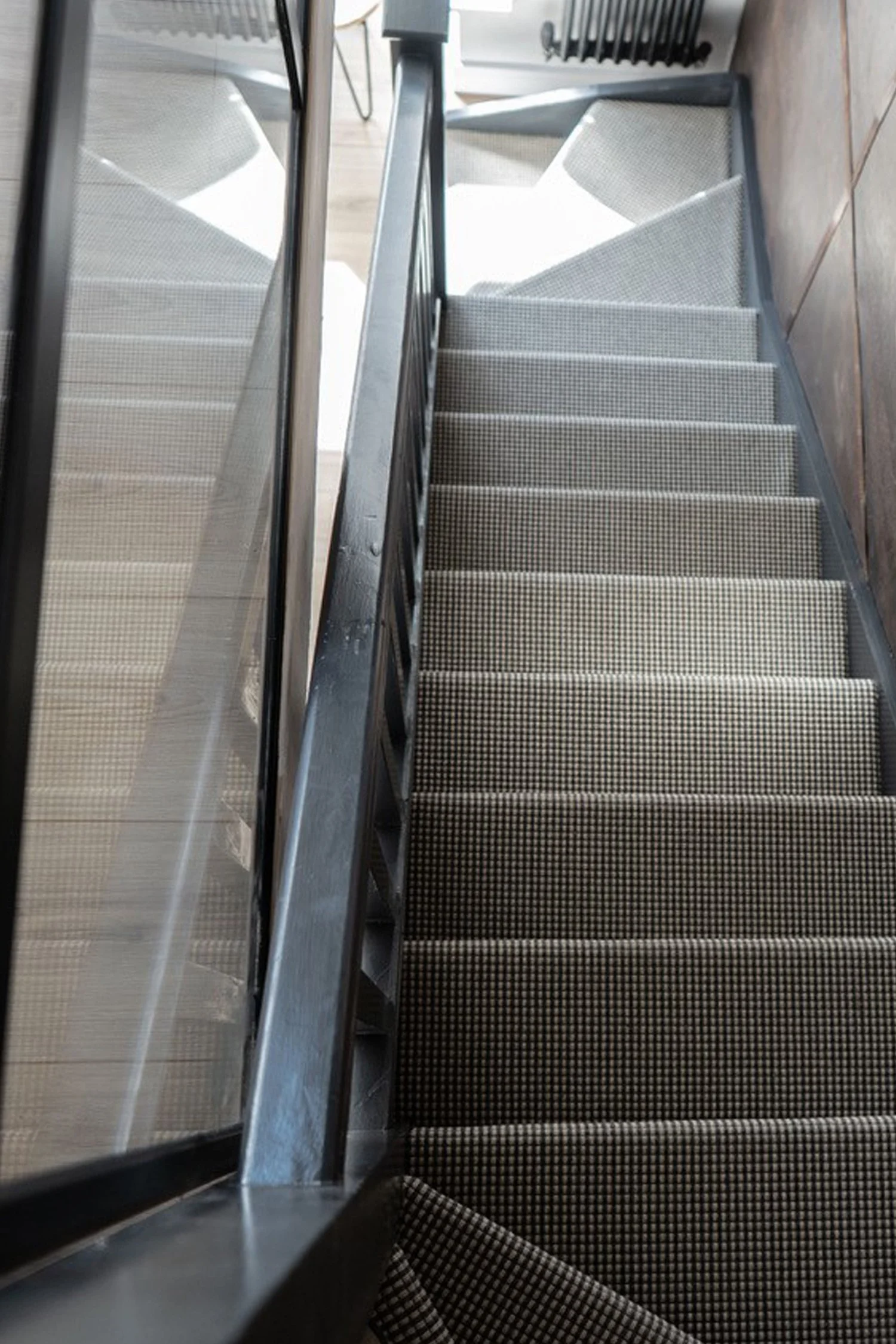 View looking down a staircase with patterned carpet, black metal railings, and glass panels on the side, adjacent to a wooden floor.