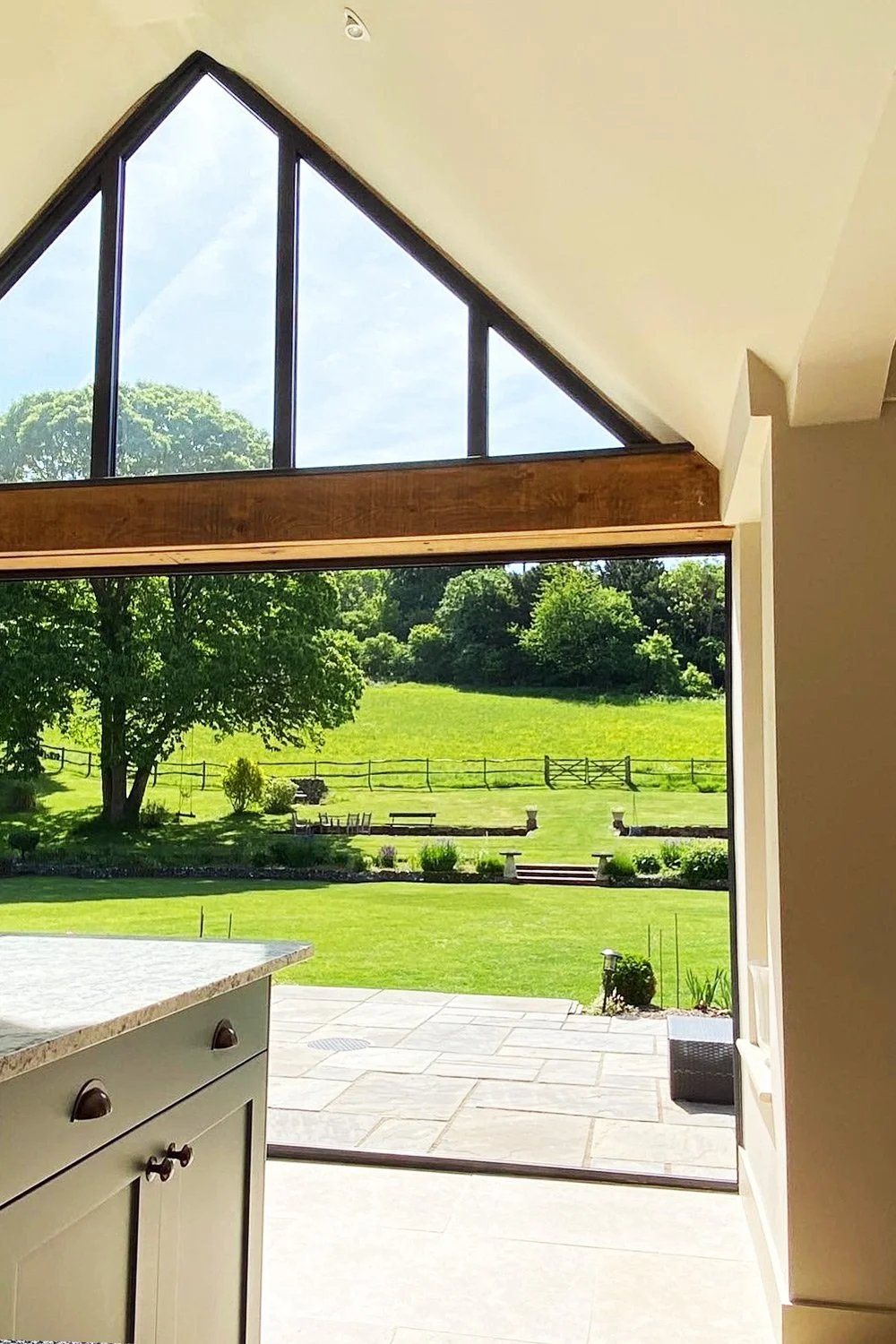 Open sliding door leading to a lush green backyard with trees, grass, and a wooden fence, viewed from inside a modern house with large triangular windows and a kitchen counter.