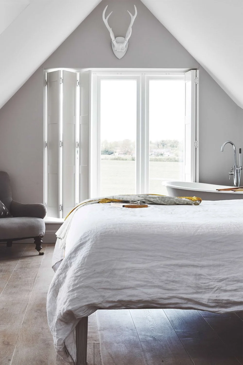 A bedroom with a white bed, gray armchair, and a balcony door with white shutters. A decorative white animal skull with antlers is mounted above the door. The room has sloped walls and wooden flooring.