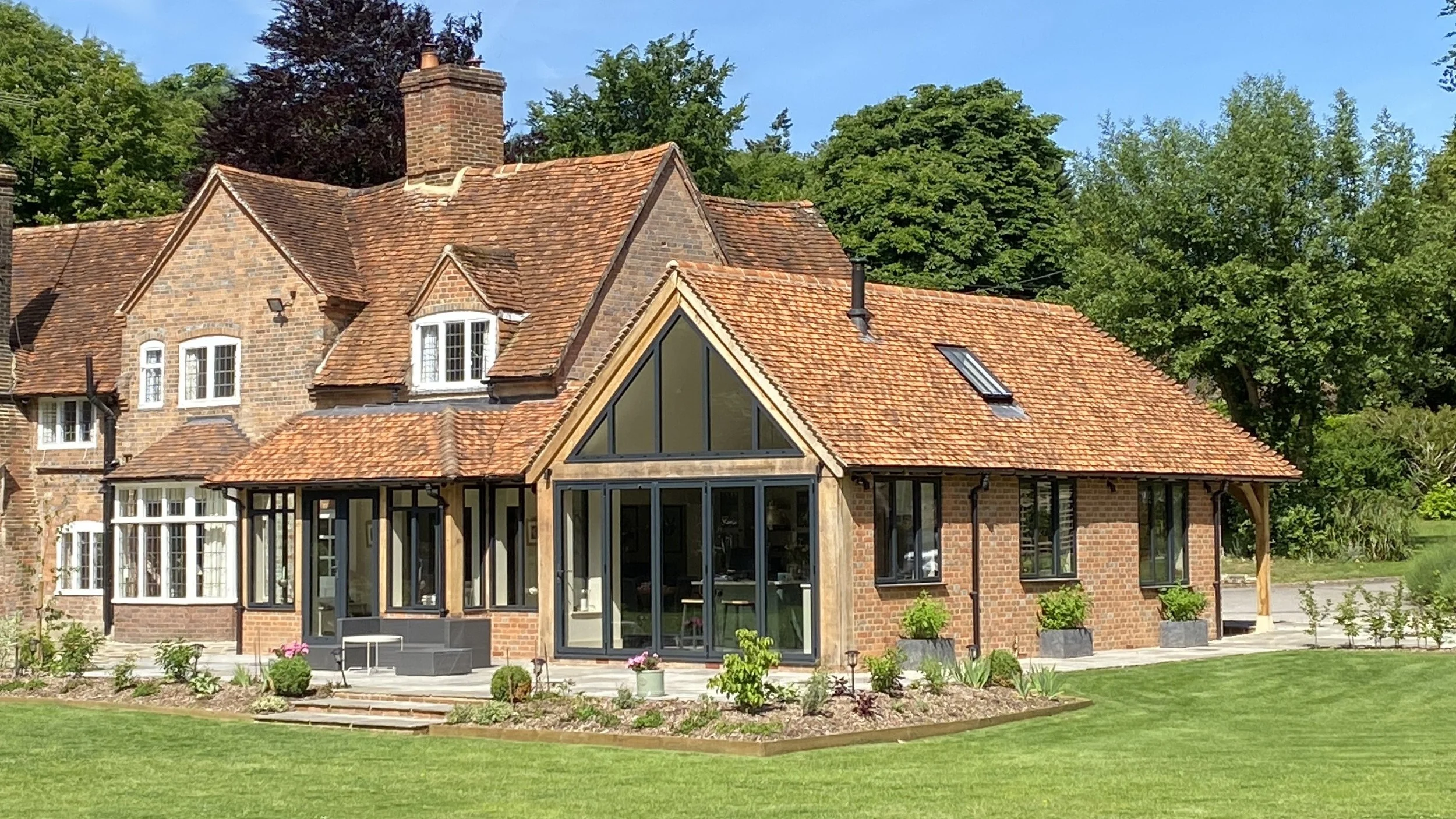 A large brick house with multiple roofs, including a glass-enclosed sunroom, surrounded by a well-maintained lawn and garden, with tall trees and a blue sky in the background.