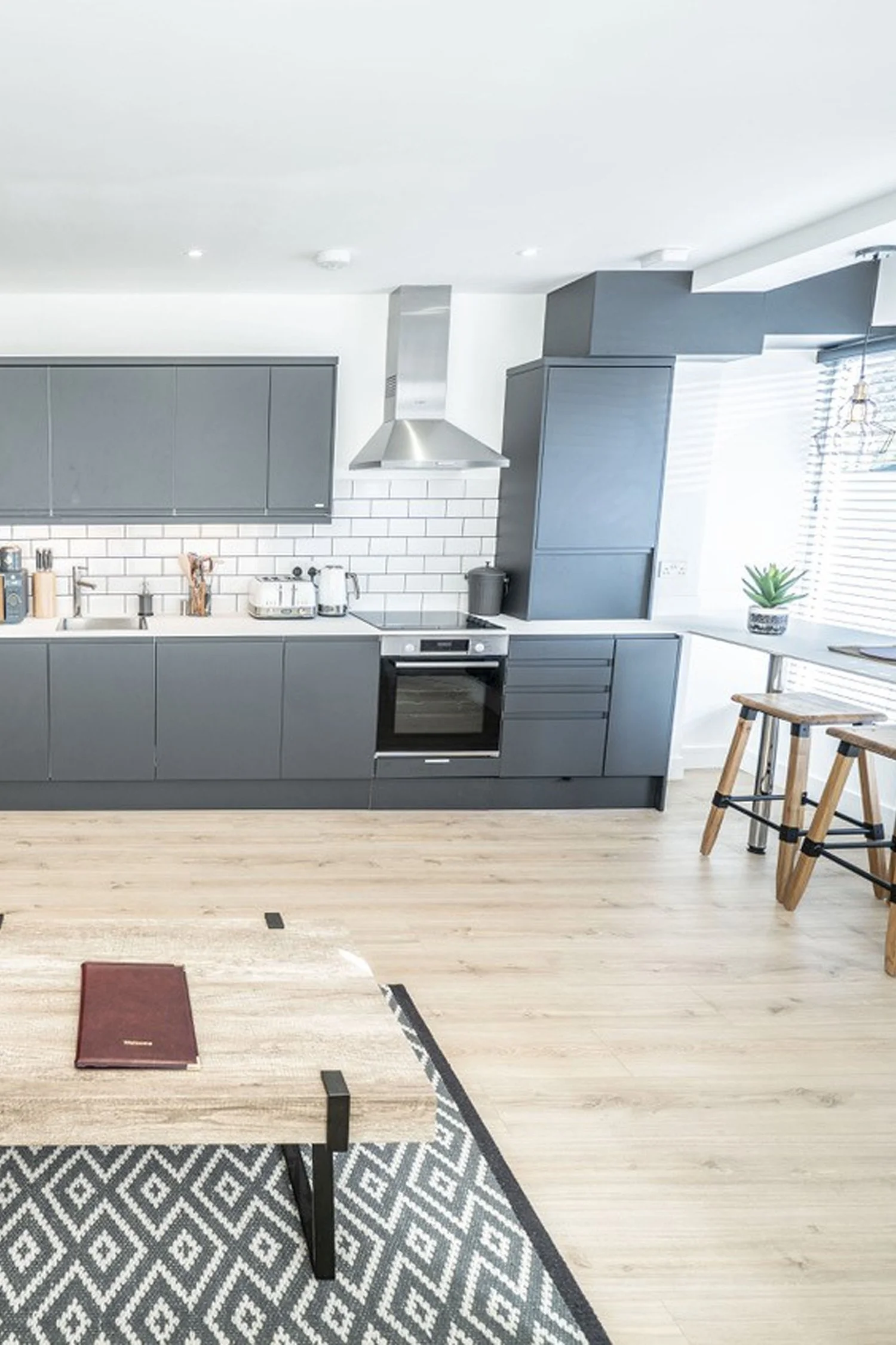 Modern kitchen with gray cabinets, white subway tile backsplash, stainless steel range hood, and light hardwood floors. There is a small table with chairs, and a window with blinds and a potted plant on the windowsill.