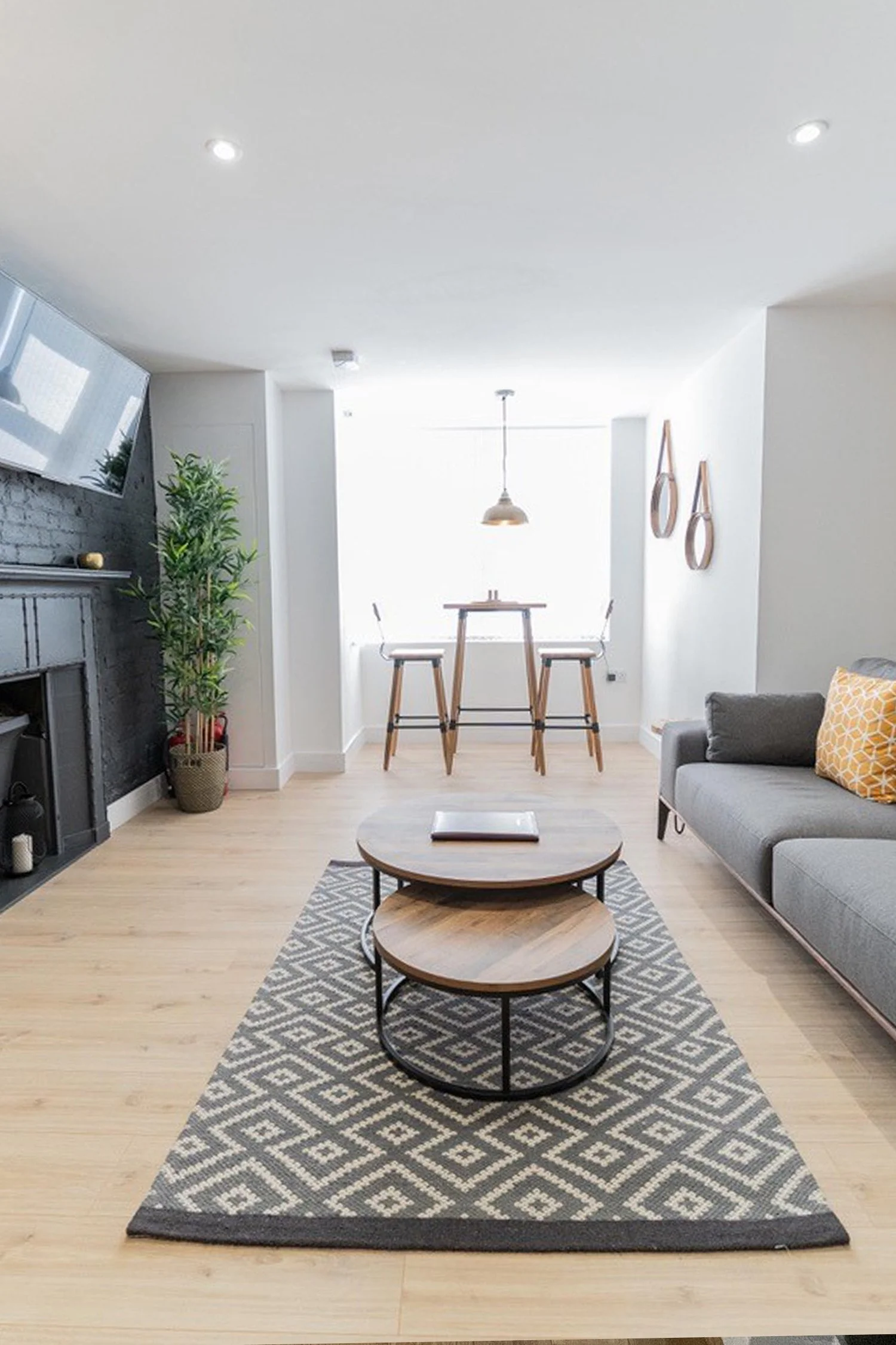Living room with gray couch, wooden coffee table, geometric patterned rug, wall-mounted television, potted plant, and dining area with bar stools and hanging pendant light.
