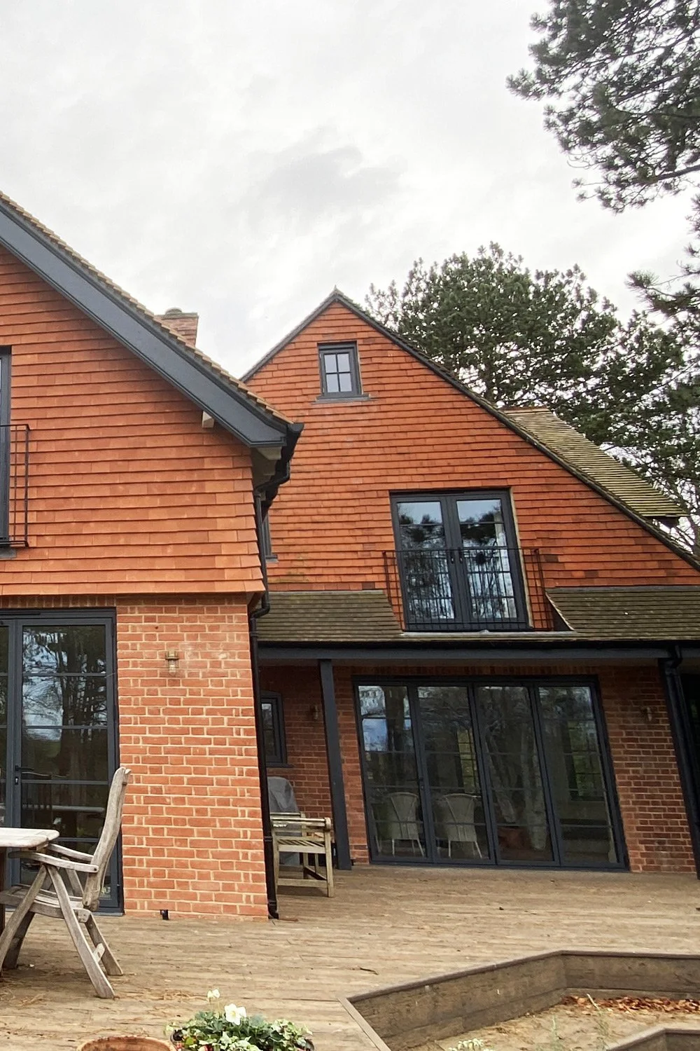 A two-story brick house with sloped roofs and large glass doors opening onto a wooden deck, surrounded by trees.