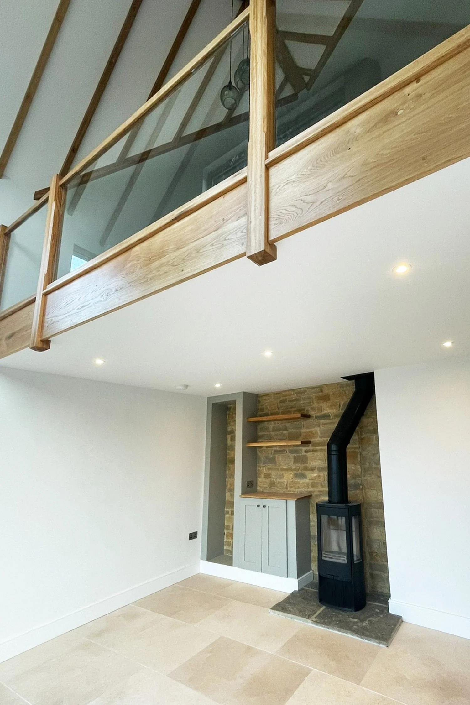 Interior of a modern home with a wooden balcony, stone accent wall, wood stove on a stone hearth, and beige tiled flooring.